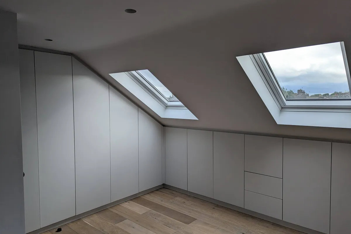 Bespoke fitted loft storage wrapping the full perimeter of a loft room, handleless light grey finish following the sloped ceiling line with integrated drawers and Velux windows