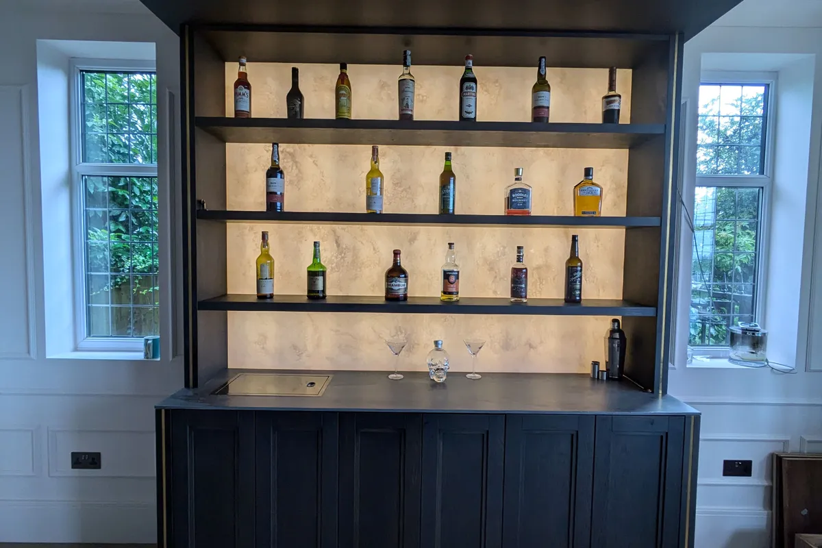Bespoke home bar in dark navy with backlit marble shelving displaying whisky bottles, panelled front, integrated sink and overhead canopy